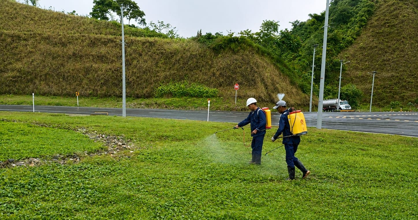Dos operarios con equipos de aspersión de espalda tratan la franja verde entre la calzada y el talud de una vía, con el corte del terreno y un camión al fondo.