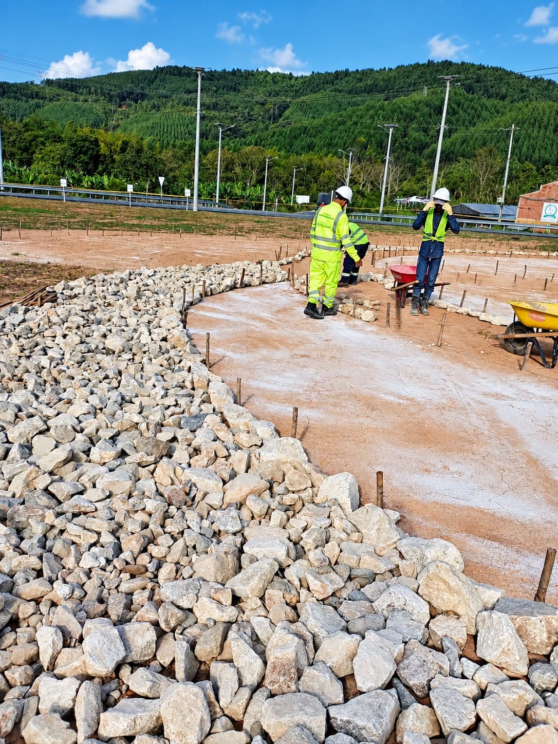 Operario con casco carga piedra a dos carretillas junto a un acopio, al borde de una vía.
