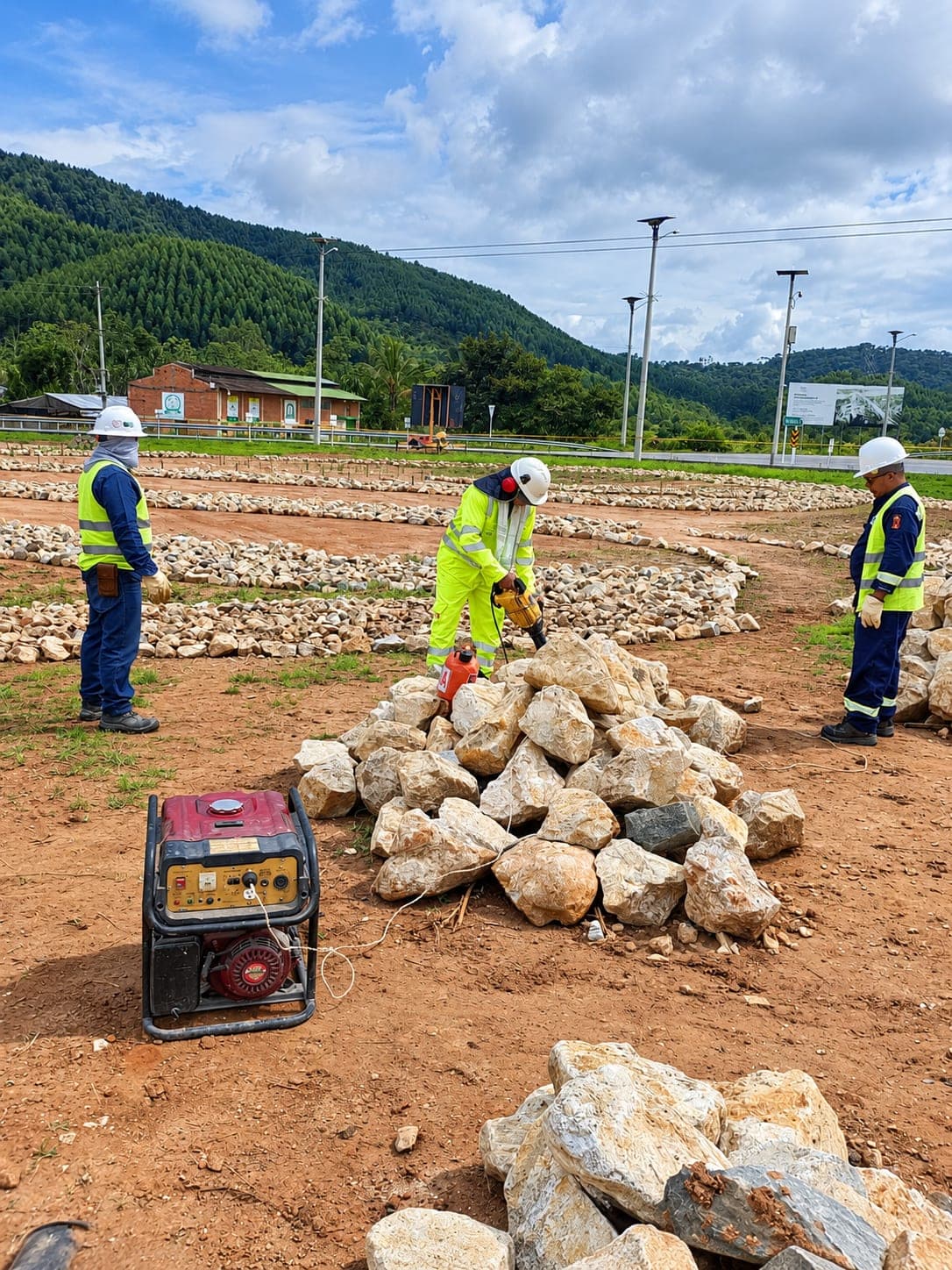 Tres operarios alrededor de un acopio de piedra; uno trabaja una roca con herramienta eléctrica alimentada por una planta portátil.